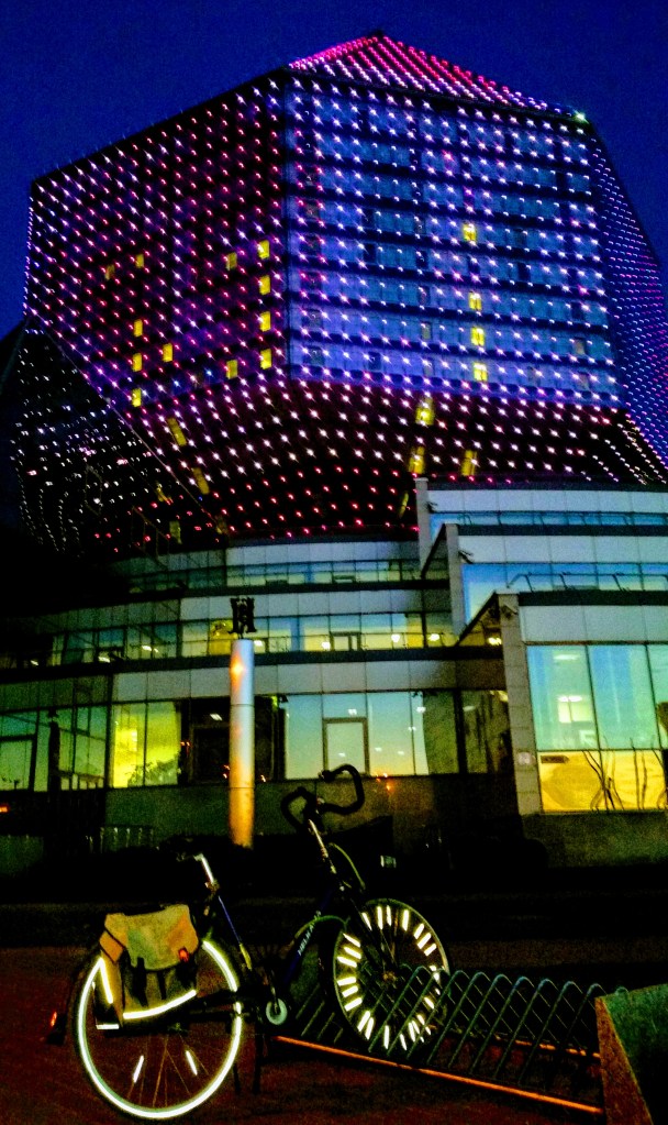 A bicycle parked in front of a diamond shaped, colorful, building in the night