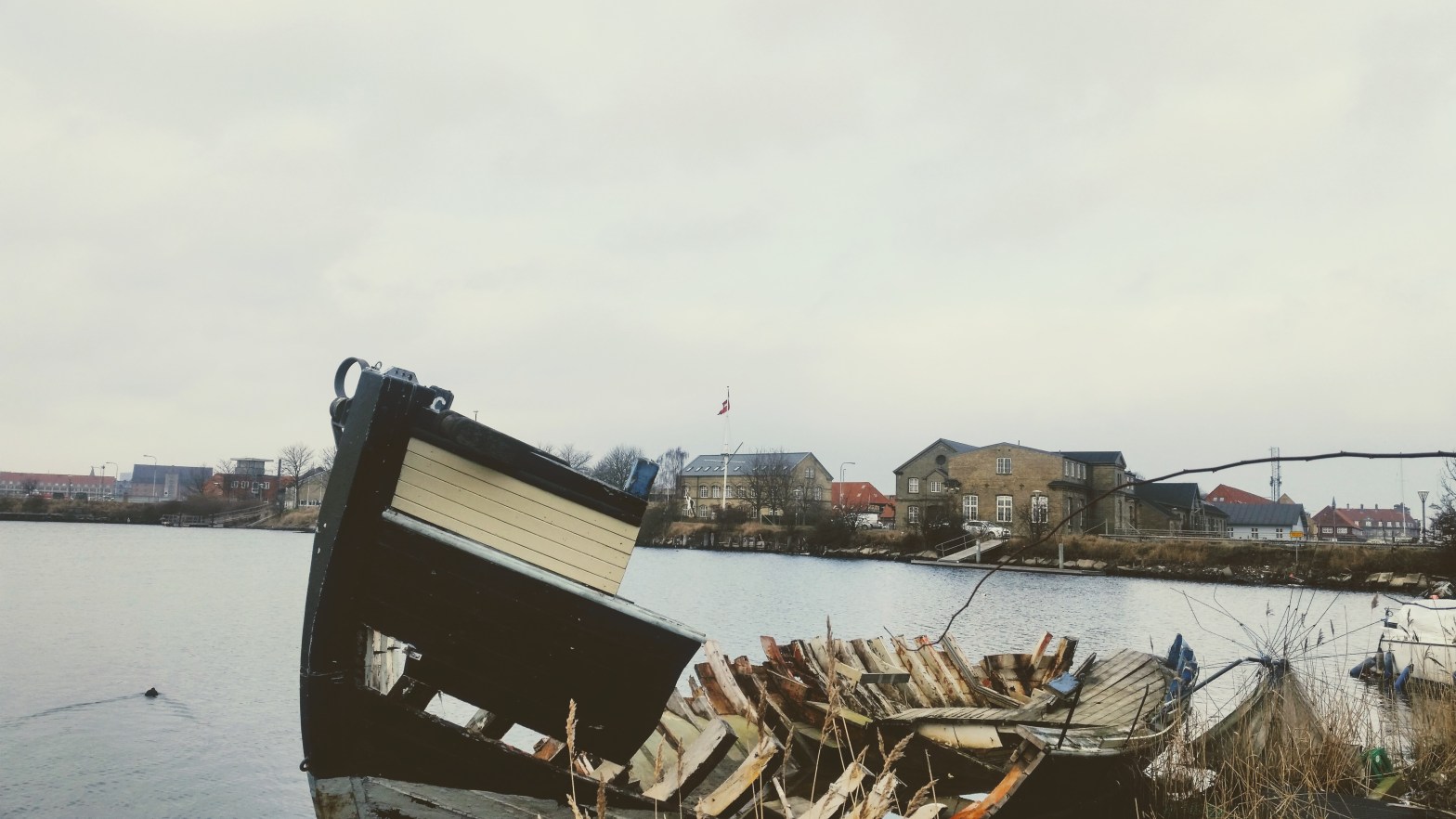 A totally broken small boat, black and white, behind which you can see some typical Danish style buildings in the outskirt of Copenhagen.